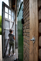 A colorful picture of a caucasian woman with blond hair standing by a big old wooden door with little windows in it in a brick wall. The woman is looking what is behind the door.