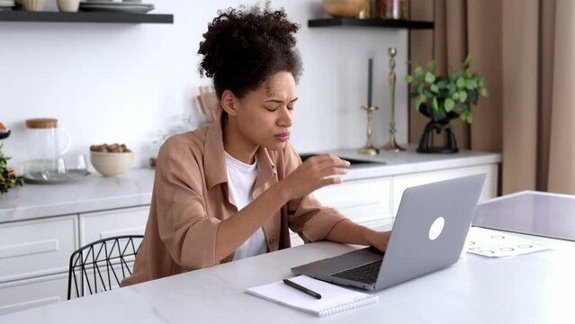 Frustrated upset african american brunette girl, freelancer, manager, sits at the table in the kitchen, uses laptop while remote work, shocked by bad news or message, suffered financial loss, despair