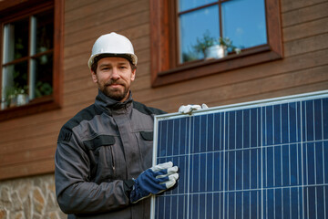 Smiling handyman solar installer carrying solar module while installing solar panel system on house.