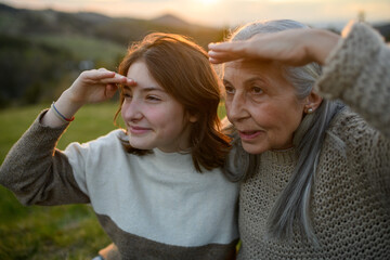 Portrait of happy senior grandmother with teenage granddaguhter sitting on grass and looking into...