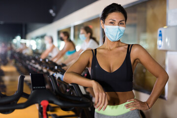 Portrait of young adult woman in protective face mask posing near stationary bike at gym