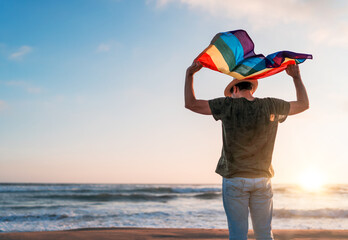person holding rainbow flag above the head, back view, LGTBQ, on the beach at sunset .