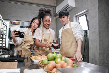 Fototapeta premium Three young students in cooking class wear aprons enjoyment while taking selfie photo with mobile phone in kitchen, smiling and laughing, preparing eggs and fruits, learn fun culinary course together.
