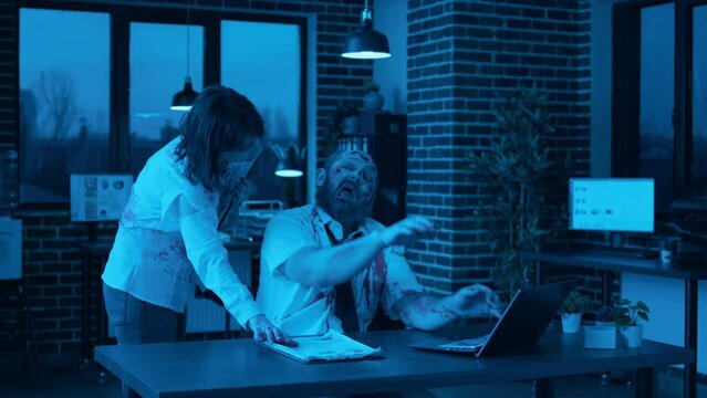 Creepy mindless zombie sitting in office while working on modern laptop at night. Spooky brain dead monster with deep and bloody scars typing on portable computer while dead walking coworker brings