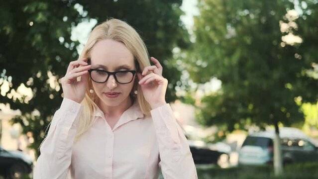 Beautiful girl in a shirt with an incorrect position of the eyes (strabismus) puts on glasses, smiles and rejoices in good vision. Against the background of green trees

