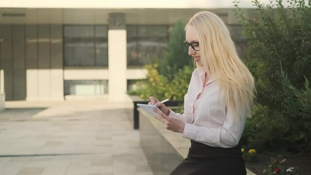 Beautiful business woman in a shirt and black skirt sits near a flower bed with a notebook and a pen in her hand. Something is recording. Side view
