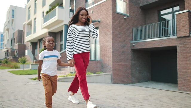 African American pretty smiling young woman walks on street in city with small happy kid boy and calling on smartphone. Mom talking on cellphone while walking with son, family concept