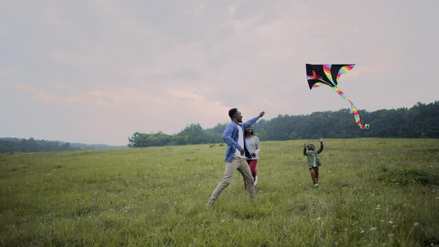 Happy Family Day, Father Mother And Little Kid Running In Field Flying A Kite Into The Sky. African American Young Cheerful Parents Play With Child In Nature Outdoor Spending Time Together With Family