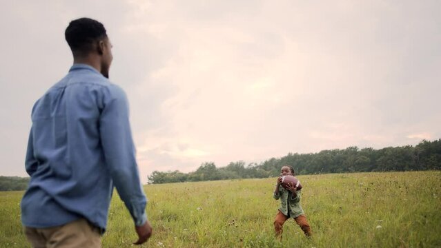 Rear of African American father playing with little son kid with ball standing in field. Cute child boy plays american football throwing and catching ball in nature. Happy childhood concept