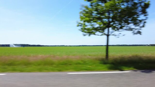 Side window view from a moving car with oncoming traffic flashing past. Travel on an asphalt road along the meadows, green fields with agricultural irrigation system. With forests and a clear blue sky