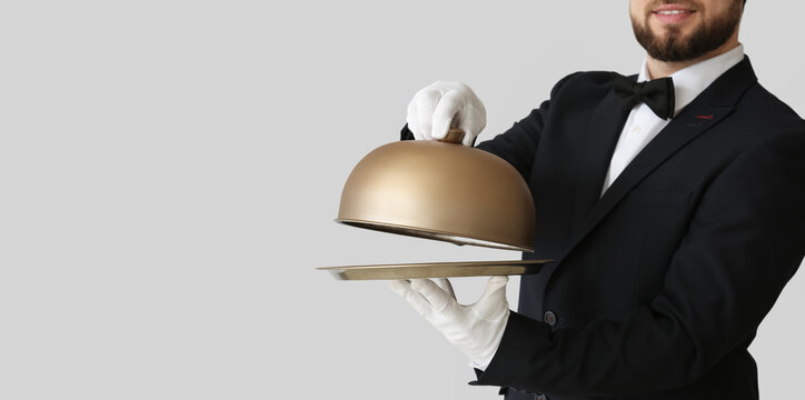 Handsome male waiter with tray and cloche on light background with space for text