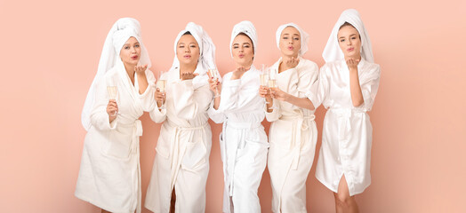 Beautiful young women in bathrobes drinking champagne and blowing kiss on color background
