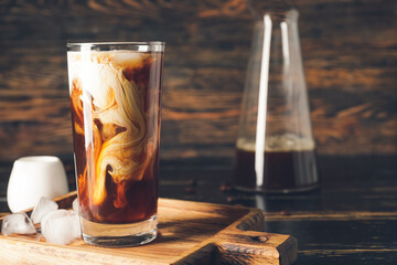 Board with glass of cold brew coffee and ice cubes on dark wooden table