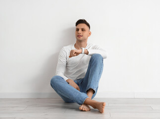 Fototapeta premium Young barefoot man with smartwatch sitting near light wall
