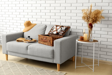 Sofa with basket for picnic, photo camera, bag and hat near white brick wall