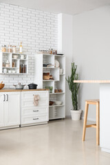 Stylish interior of light kitchen with modern counters and shelf unit near white brick wall