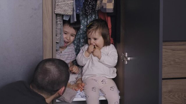 Dad playing hide-and-seek with his children around the house. He opens the closet door to find the children hidden there. Sitting and smiling in dad's wardrobe