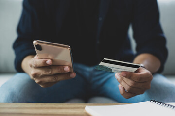 Young man sitting on sofa holding credit card while using smart phone for shopping online at home. He likes to order groceries online. He thought it was convenient and easy to pay by credit card.