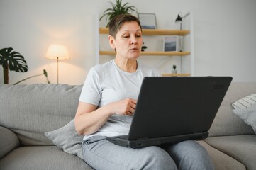 Older mature woman using wireless laptop apps browsing internet sit on sofa, smiling middle aged grandmother working distantly on computer surfing web communicating online looking at screen at home.