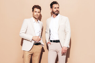 Portrait of two handsome confident stylish hipster lambersexual models. Sexy modern men dressed in white elegant suit. Fashion male posing in studio near beige wall