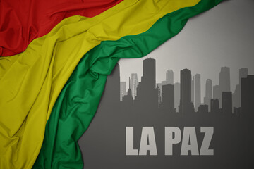 abstract silhouette of the city with text La Paz near waving national flag of bolivia on a gray background.