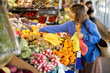 Woman choosing fresh fruits at the local market, shopping with reusable mesh bag. Sustainability...