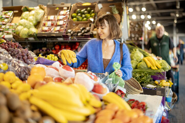 Young cheerful woman choosing fresh fruits at local market, shopping with reusable mesh bag. Sustainability and organic local food concept