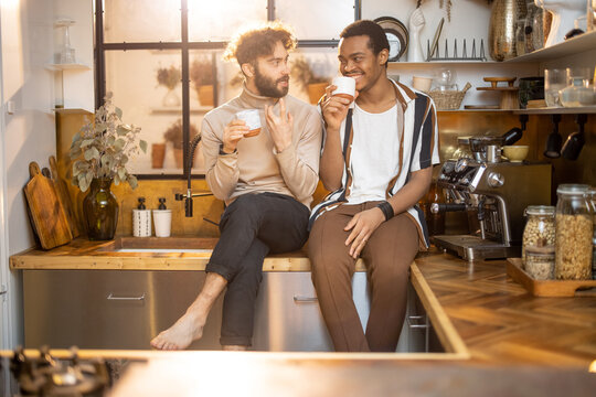 Two guys of different ethnicity having warm conversation while sitting with coffee on kitchen at home. Concept of close male friendship or relationship as gay. Caucasian and hispanic man together