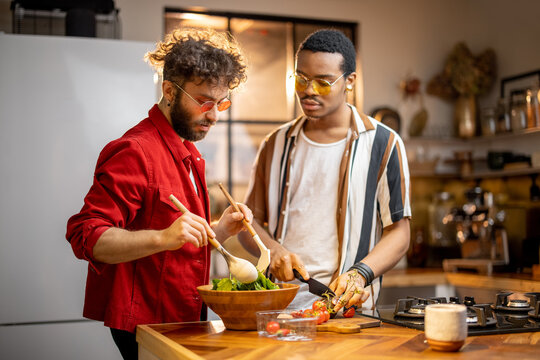 Two brightly dressed guys having fun while making salad together on kitchen. Concept of gay couples and everyday life at home. Caucasian and hispanic man cooking healthy food