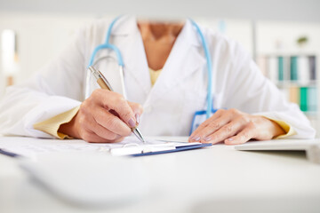Close-up of unrecognizable doctor sitting at table and making notes in medical record while working with medical papers