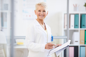 Portrait of positive confident female doctor in lab coat standing in office and working with medical files