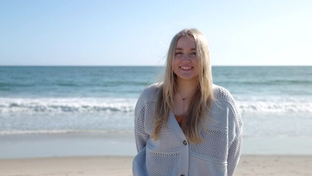 Portrait of a woman chilling at the beach. Pretty young lady smiles, while ocean breeze shaking her blonde hair. High quality 4k footage
