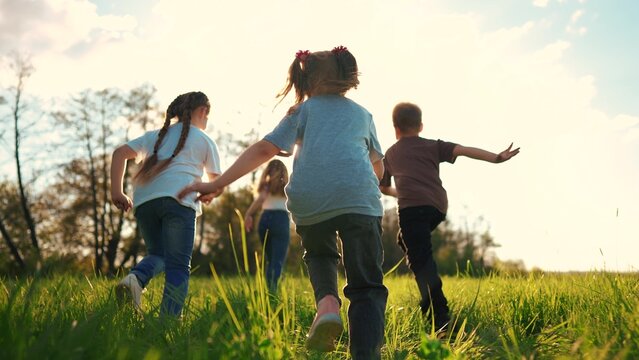 Kids Run In The Park. A Large Group Of A Team Of Children Running Back View Sunlight In The Summer On The Grass In The Park Camera Movement. People In The Park Happy Family Kid Lifestyle Dream Concept