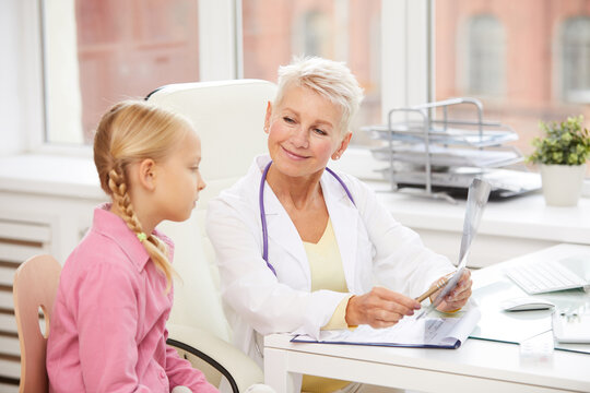 Content friendly mature female pediatrician pointing at x-ray image while explaining reason of pain to little girl in childrens healthcare clinic