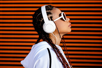 Young African American woman with braided hair wearing urban sportswear with headphones and sunglasses on an orange background