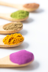 Multicolored superfood powders in wooden spoons: sweet potato, matcha, spirulina, hemp, turmeric, carob on a white background. Selective focus.