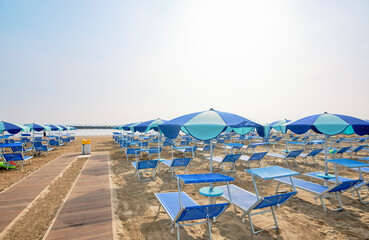 Boardwalk on the beach along a row of colorful umbrellas and sunbeds leading to the sea on a sunny summer day on the shore of the Adriatic Sea, Italy. Early morning sun on the beach.