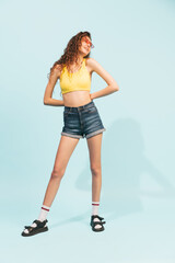 Full-length portrait of young cheerful girl posing in casual outfit, shorts and top isolated over light blue studio background