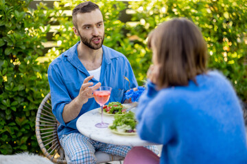 Young stylish couple have conversation while sitting by the round coffee table at their beautiful garden. Young man and woman have romantic evening outdoors