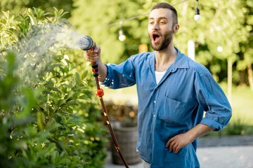 Rolgordijnen Handsome guy watering green hedge at garden. Husband take care of bushes at backyard of his house on summer time © rh2010