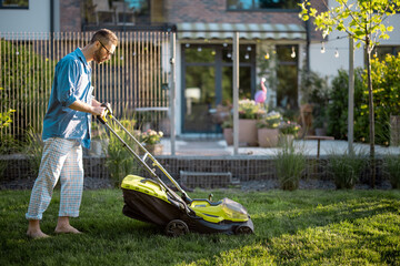 Man mows the lawn with lawn mower at backyard of his house. Husband takes care of garden on summer evening. Modern electric wireless garden equipment - obrazy, fototapety, plakaty