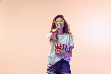 Portrait of young girl in casual outfit posing with popcorn basket isolated over pink studio background in neon light. Comedy film