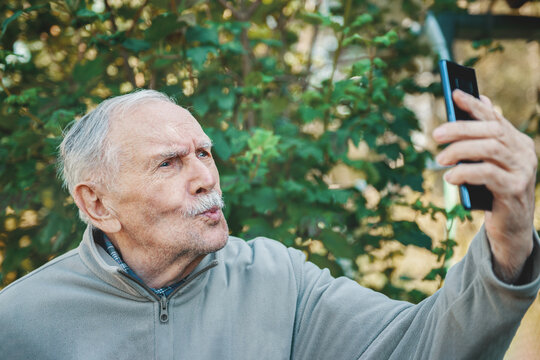 grey man takes a selfie from his mobile phone to post it on social media for communication. A man of 88 years old communicates with his grandchildren on a smartphone, active old age