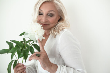 Portrait of a beautiful woman aged 60, dressed in a white shirt, holding flowers against a wall