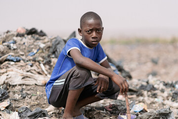 Young African waste picker boy sitting crouched in a landfill surrounded by plastic garbage,...