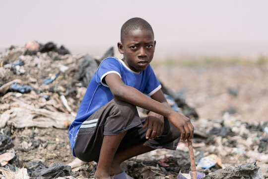 Young African waste picker boy sitting crouched in a landfill surrounded by plastic garbage, looking at the camera with a frown; symbol of stolen childhood and child labour