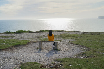 A woman in a yellow jacket sits on a bench on top of a mountain and looks at the sea and huge rocks at sunset.