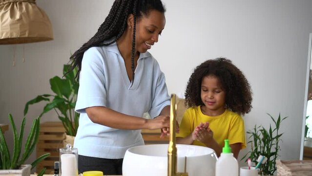 Mom, daughter are washing hands and talking while standing in home bathroom spbd. Close view of american african woman, girl clean dirty skin and use foam, talk with smiles and stand in light interior