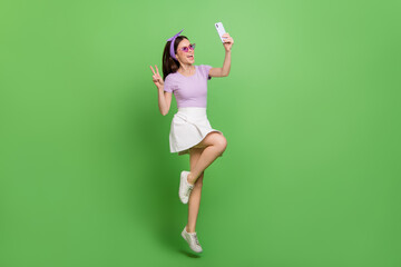 Full size funny photo of cool brunette lady jump do selfie wear head band glasses t-shirt skirt sneakers isolated on green background