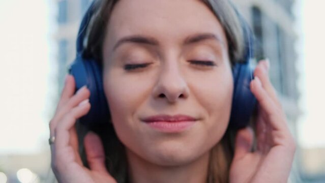 Blue eyes happy young woman in headphones raises head and looks at the camera outdoors in city street having fun alone. Joyful attractive blonde carefree woman listening to music in urban town.